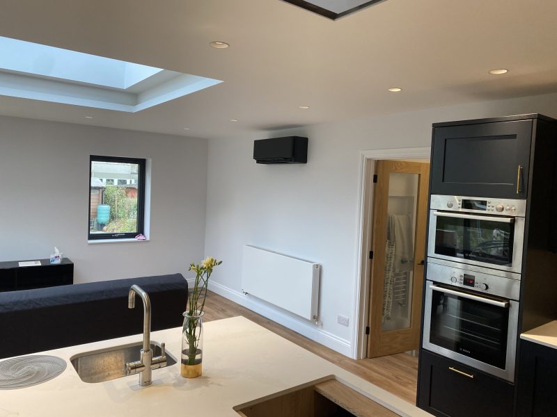 Modern kitchen and living area with a skylight, white walls, black appliances, and wooden flooring. There's a sink with flowers on the counter and a sofa near a window and door.