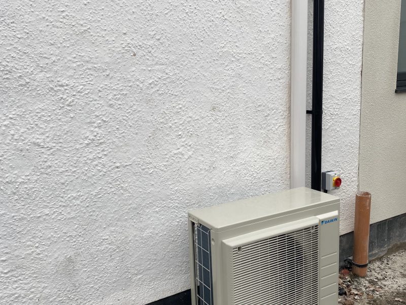 A white outdoor air conditioning unit is installed against a textured white wall. There are pipes leading from the unit into the wall, and dry leaves are scattered on the ground below.