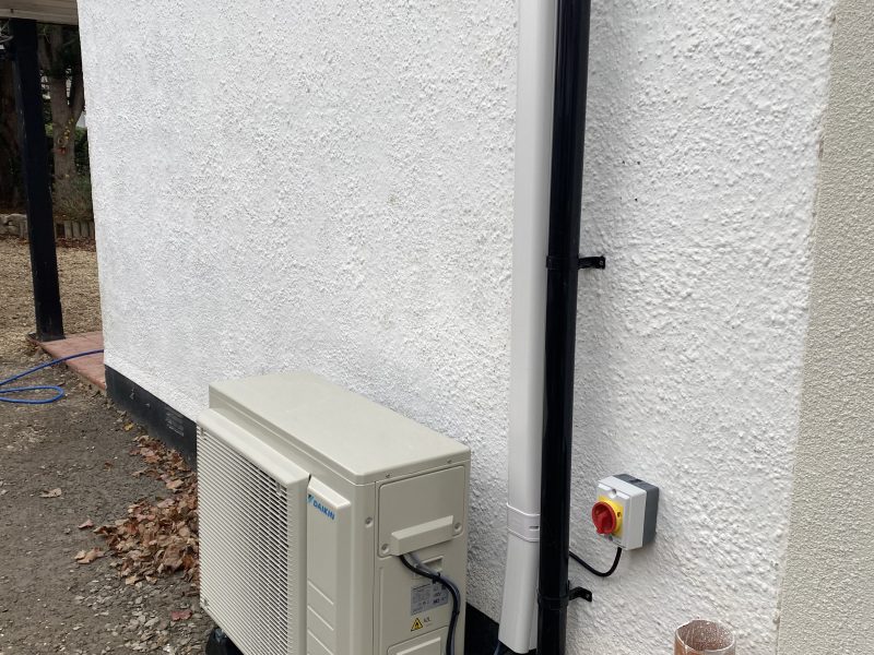 Outdoor air conditioning unit installed on a white wall, with cables and pipes connected. A red emergency stop button is mounted on the wall nearby.