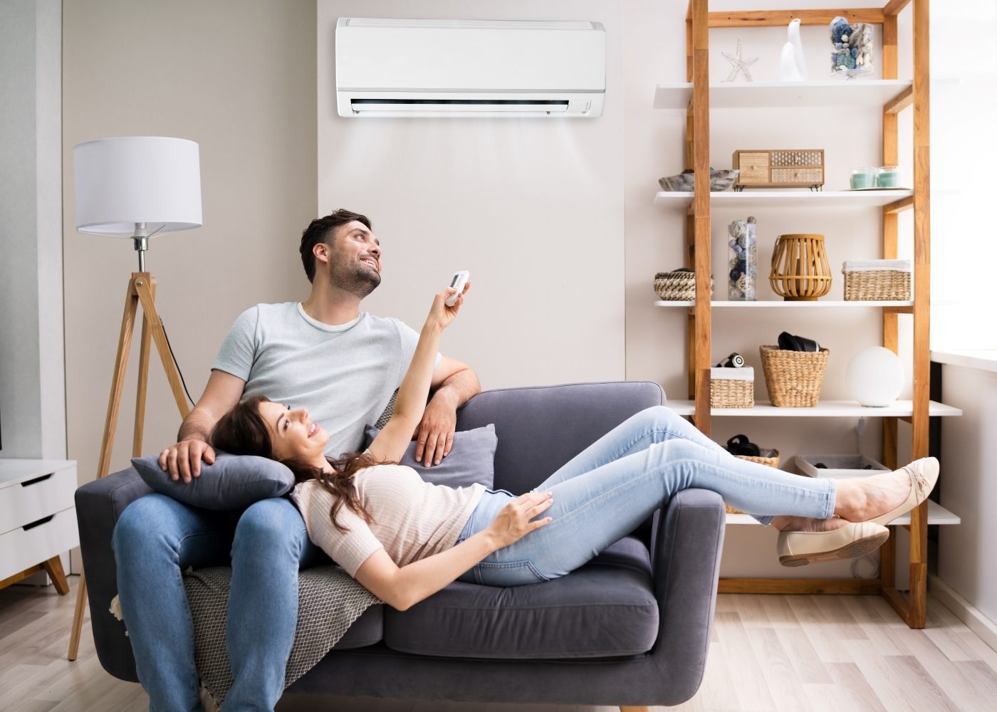 A couple relaxes on a sofa under an air conditioner. The woman holds a remote, and the room includes a shelf with various items and a floor lamp.