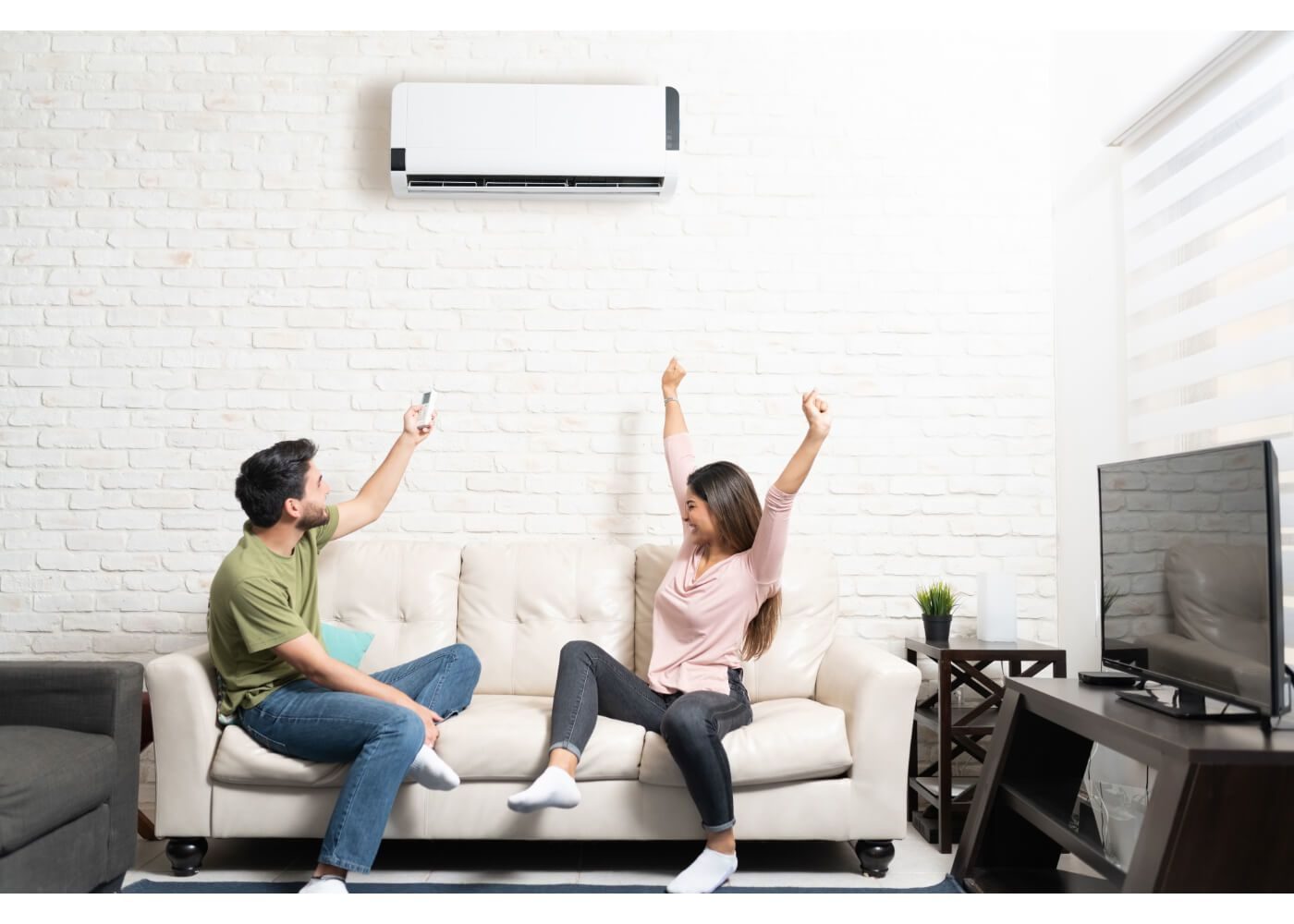 A man and woman sit on a sofa, adjusting an air conditioner with a remote. The woman raises her arms in a relaxed manner.