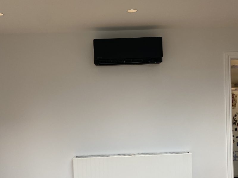 A white wall with a black air conditioning unit mounted above a white radiator. Ceiling spotlights are visible, and there is a doorway to the right, partially showing another room.