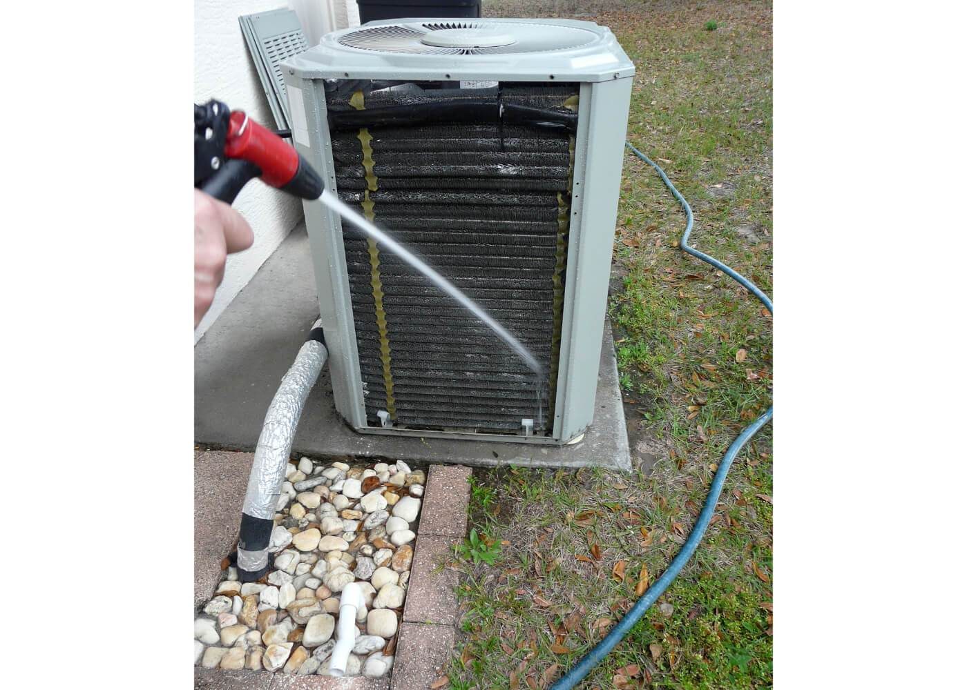 Person cleaning an outdoor air conditioning unit with a hose, surrounded by grass and rocks.