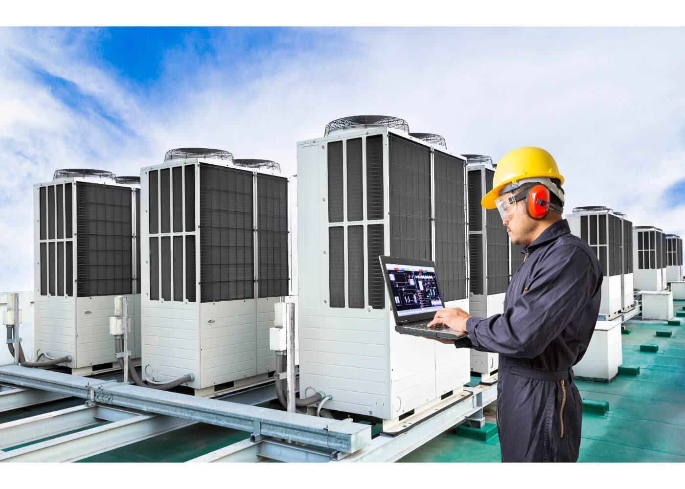 A technician in safety gear uses a laptop to monitor large air conditioning units on a rooftop.