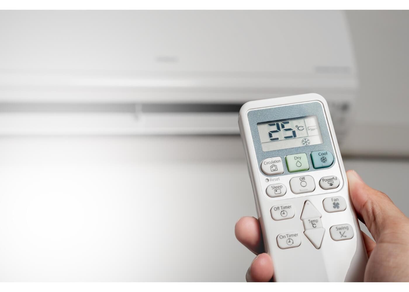 A hand holds a remote control in front of a wall-mounted air conditioning unit, displaying a temperature setting of 22 degrees Celsius.