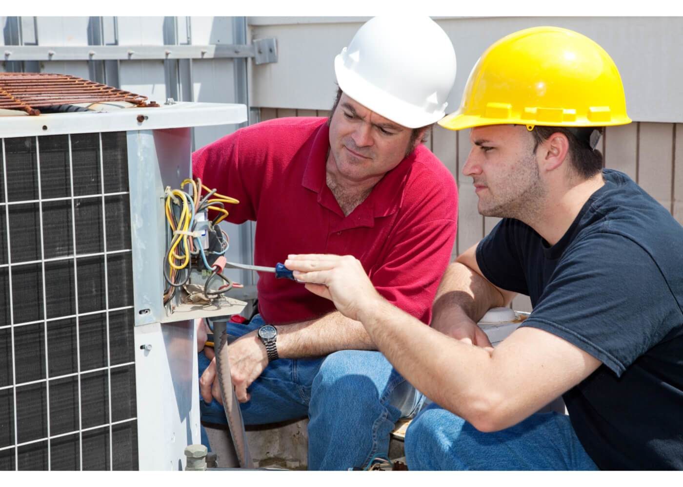 Two technicians in hard hats are inspecting and repairing an outdoor air conditioning unit. One holds a multimeter, while the other supervises.