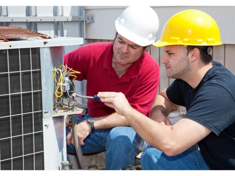 Two men wearing hard hats work on an outdoor air conditioning unit. One uses a screwdriver while the other observes.