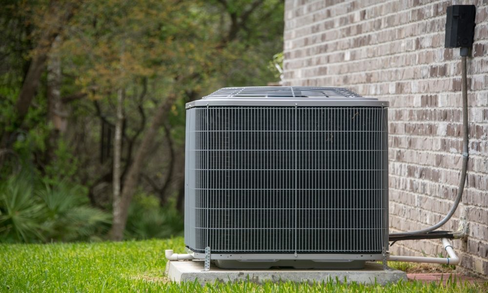 Outdoor air conditioning unit installed next to a brick wall, surrounded by grass and trees in the background.