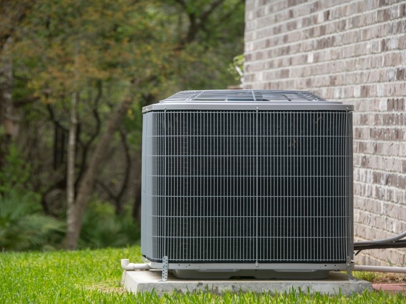 Outdoor air conditioning unit installed next to a brick wall, surrounded by grass and trees in the background.