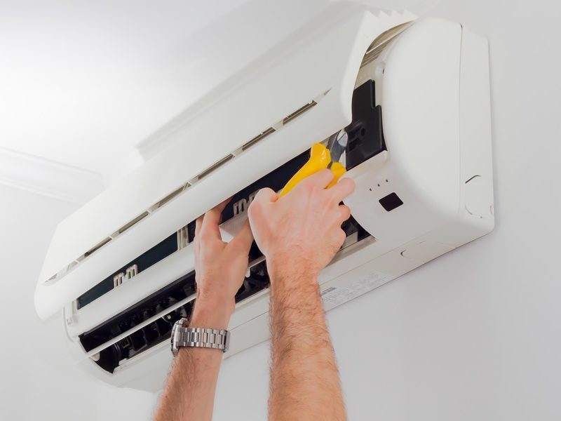 Hands wearing a wristwatch repair a wall-mounted air conditioning unit using a yellow tool.