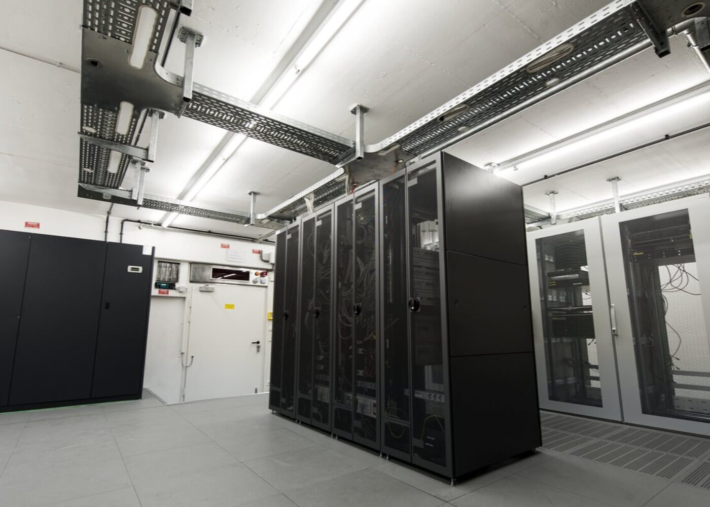 A server room with multiple black server racks, cables organised above, and fluorescent lighting on the ceiling.