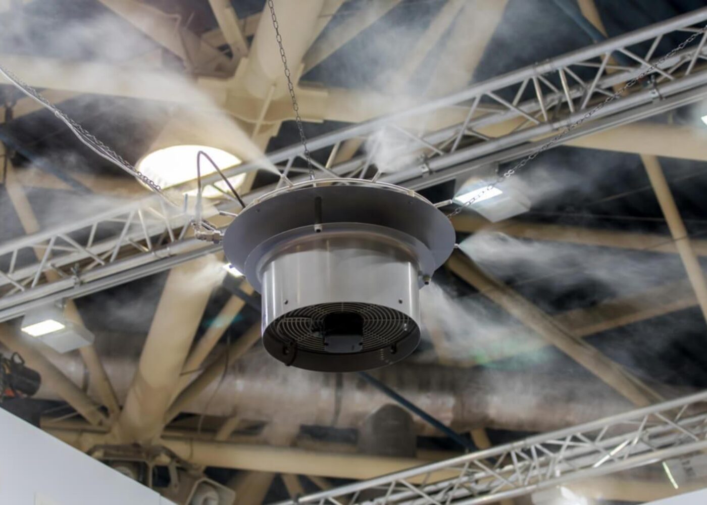 Ceiling-mounted fan emitting mist in an industrial setting with visible trusses and lights.