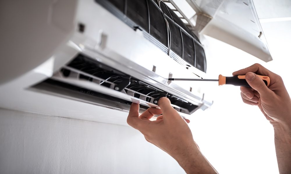 Hands using a screwdriver to repair or maintain an air conditioning unit mounted on a wall.