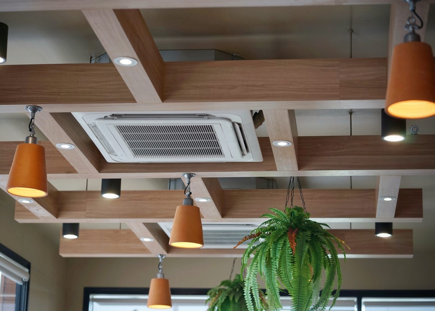 A wooden ceiling with recessed lights, an air conditioning unit, and hanging plants, including ferns and orange pendant lights.