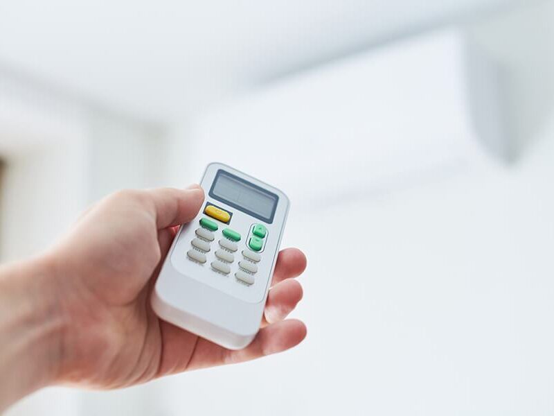 A hand holding a remote control, pointing it towards an air conditioning unit mounted on the wall.