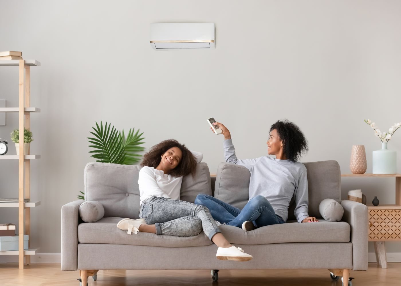 Two people are relaxing on a sofa. One holds a remote control, pointing at an air conditioner on the wall.