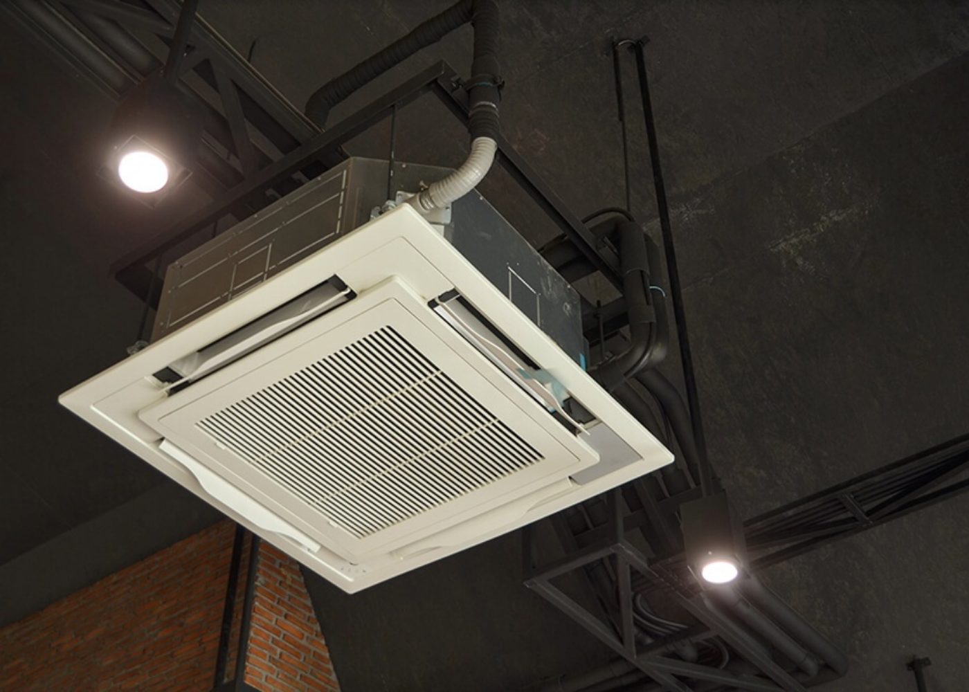 Ceiling-mounted air conditioning unit with a vent, surrounded by exposed ductwork and lighting fixtures.