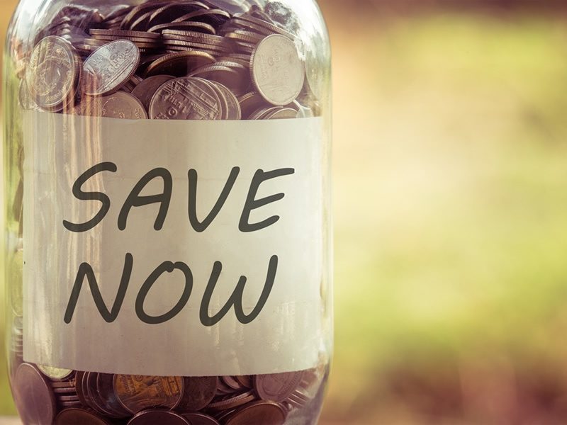 A glass jar filled with coins, labeled "SAVE NOW," against a blurred background.