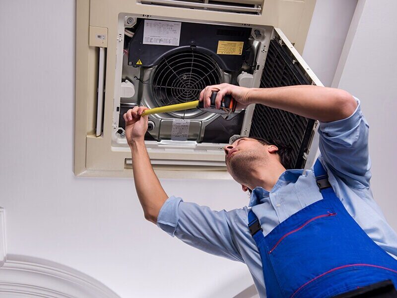 Person in blue overalls measures a ceiling-mounted air conditioning unit with a tape measure.