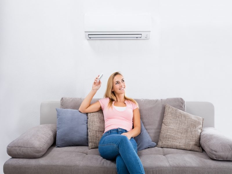 A woman sitting on a gray sofa points a remote control at a wall-mounted air conditioner.