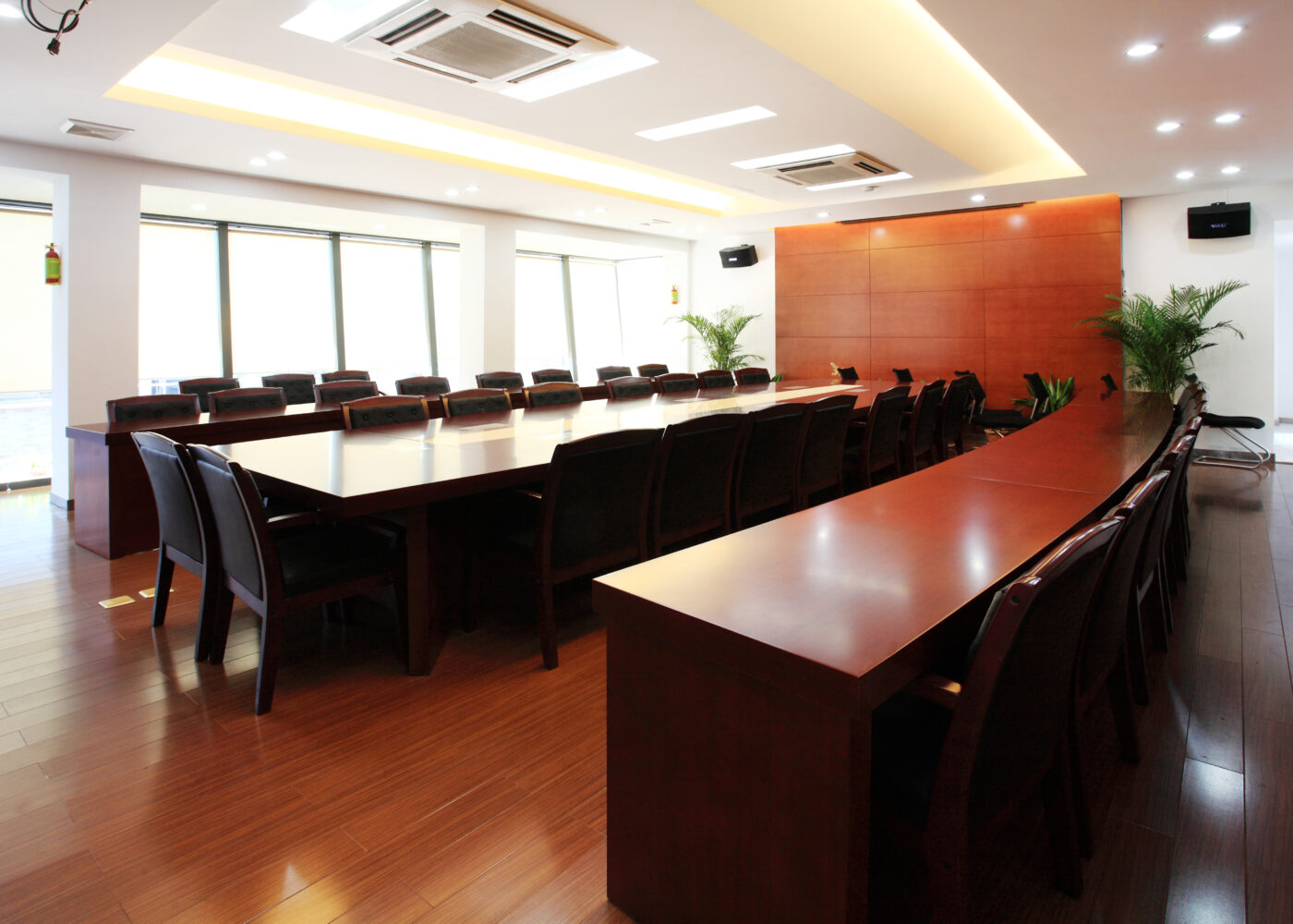 A modern conference room with a long wooden table, black chairs, hardwood flooring, large windows, and potted plants.