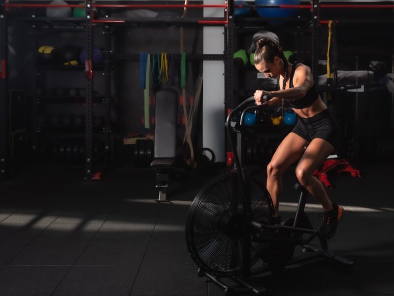 Person exercising on an air bike in a dimly lit gym, surrounded by various fitness equipment.