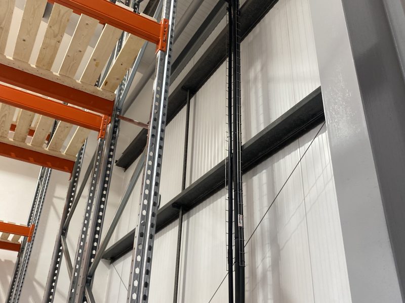 Industrial warehouse interior with metal shelving and black piping along a white wall. Wooden panels are visible on the shelves. A yellow ladder is partially visible at the bottom.