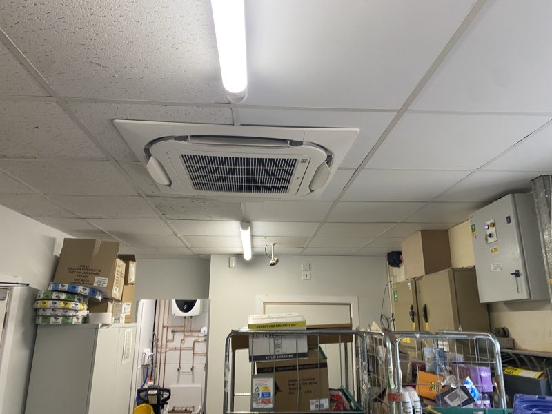 A storage room with a ceiling-mounted air conditioning unit. The room contains shelves, boxes, and electrical panels. Fluorescent lights illuminate the space.