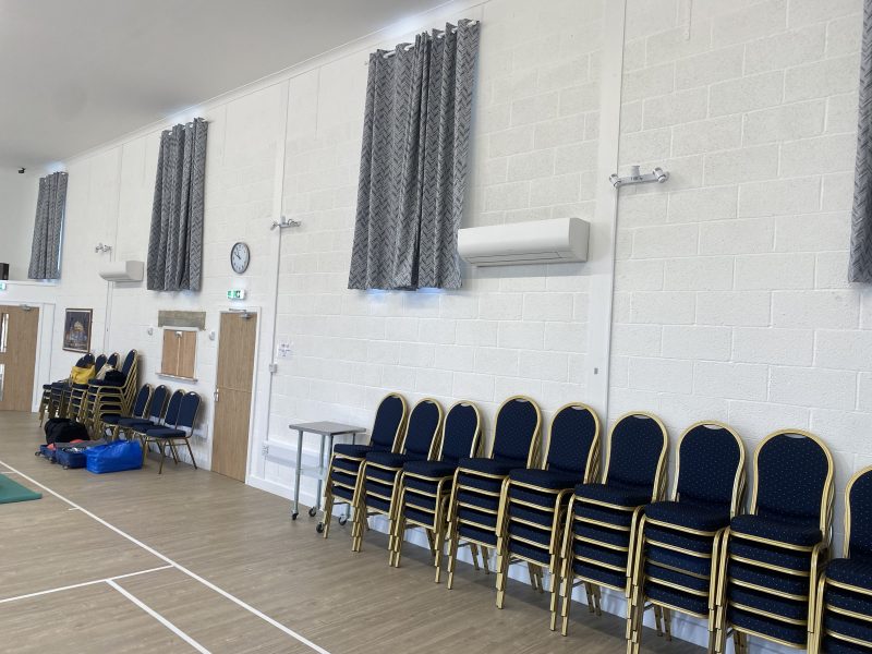 A room with stacked chairs, white brick walls, grey curtains, and a clock. Doors and fluorescent lights are on the walls. A blue mat and various objects are on the floor.