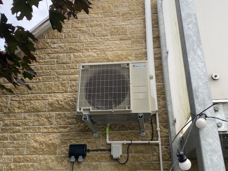 Wall-mounted outdoor air conditioning unit on a brick building, with surrounding pipes and wires. Tree branches partially visible in the upper left corner.