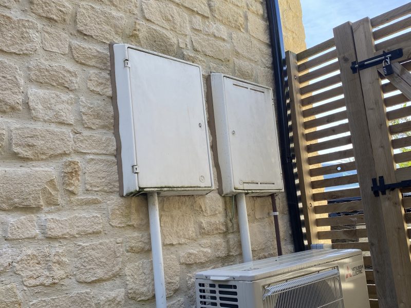 Two electrical boxes and a central air conditioning unit are mounted on a brick wall next to a wooden privacy screen.