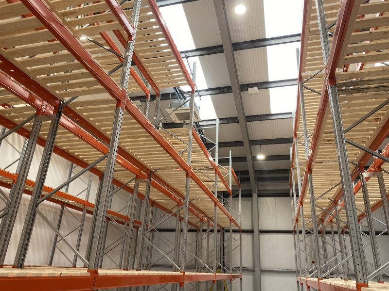 Empty industrial warehouse shelving with orange metal frames and wooden shelves, under a high ceiling with skylights.