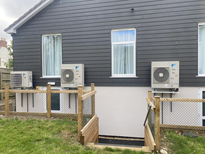 A modern building exterior with three air conditioning units mounted on a dark grey wall above a white section, surrounded by a small fenced area leading to a lower level.