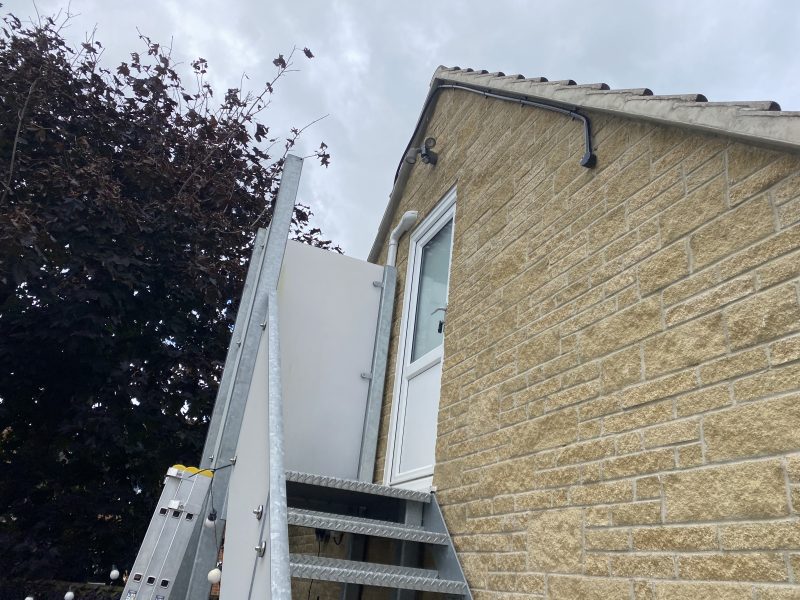 Exterior metal staircase leading to a white door on a stone building, with a dark tree on the left and a cloudy sky above.