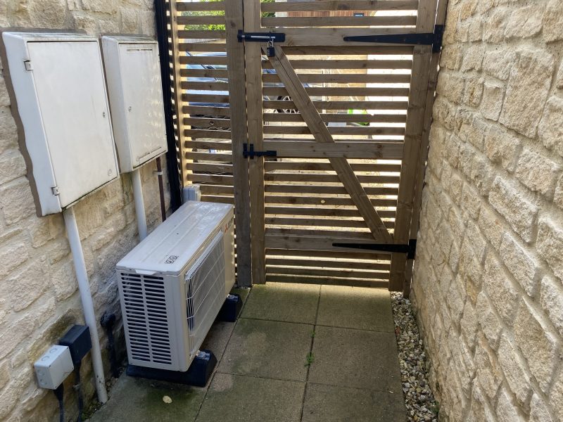 A wooden gate with horizontal slats is set in a stone wall. An air conditioning unit is installed on the ground beside the wall. Two utility boxes are mounted on the wall adjacent to the gate.