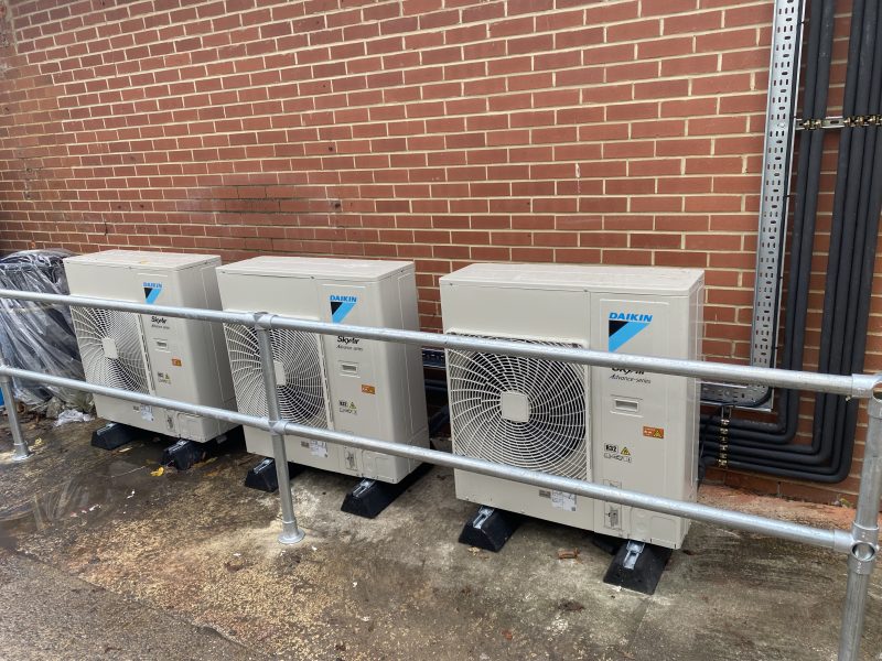 Three air conditioning units are aligned against a brick wall, enclosed by a metal railing, with piping extending vertically along the bricks.