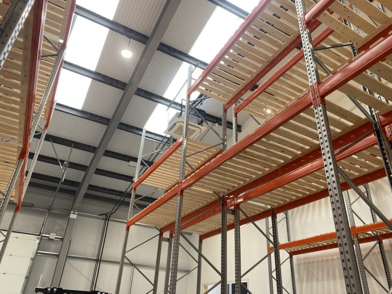 Empty industrial shelving units under a high ceiling in a warehouse with metal beams and overhead lights.