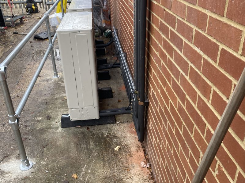 Four outdoor air conditioning units are installed along a red brick wall on a concrete surface, with a metal railing parallel to the wall.