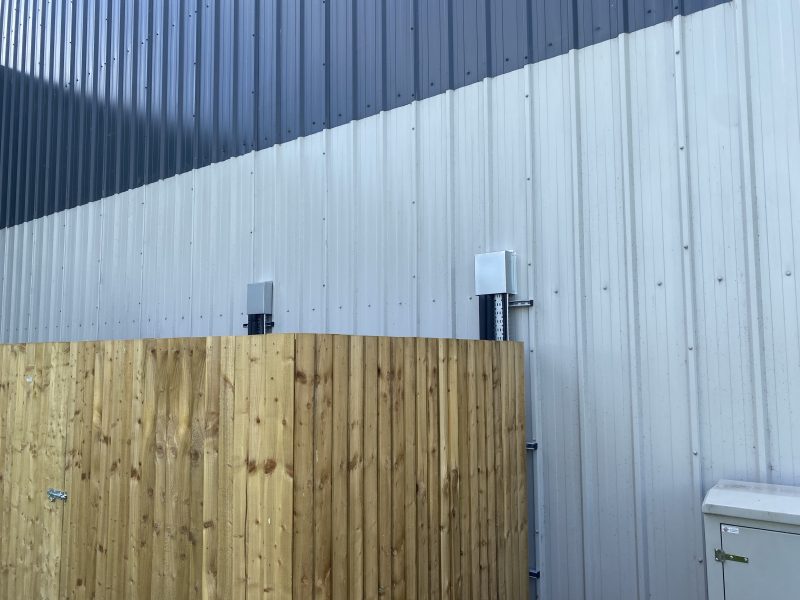 A wooden fence stands in front of a metal-clad building exterior with antennas attached to the wall.