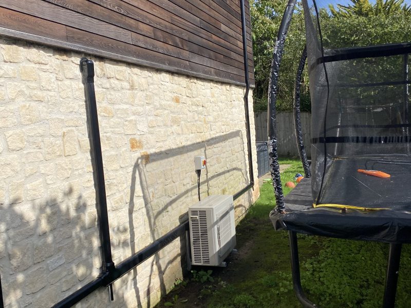 A trampoline is set up on grass beside a house with stone and wooden exterior walls. An air conditioning unit and pipes are mounted on the wall. Shadow of a fence is visible.