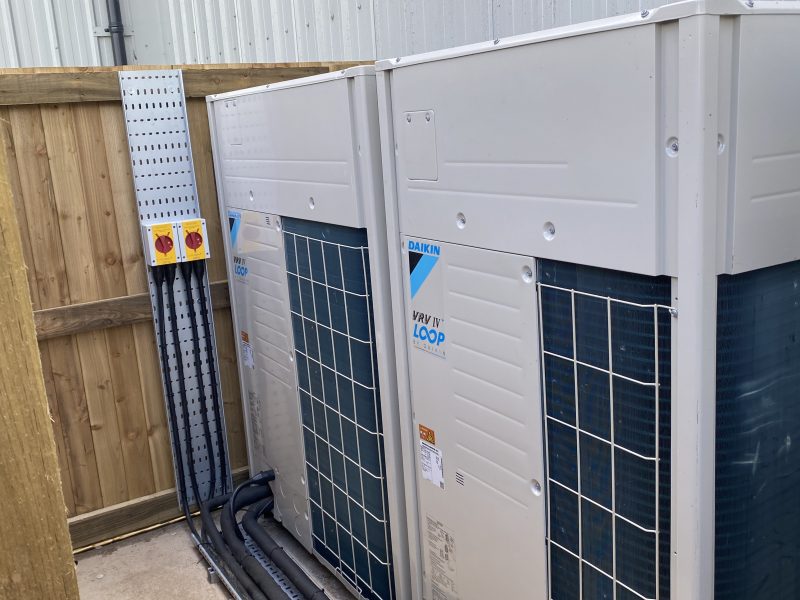 Two large air conditioning units installed outside next to a wooden fence and metal siding, with control switches mounted on the wall.