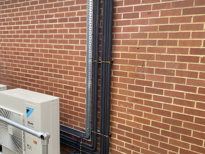 A brick wall with several black pipes vertically attached, connected to a white outdoor air conditioning unit. A metal railing is in the foreground.