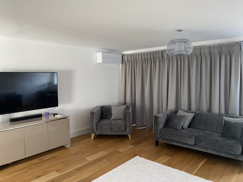 A modern living room with a wall-mounted TV, gray sofas, a matching armchair, a light wood floor, a white rug, and gray curtains. A ceiling light fixture hangs above.