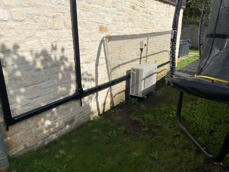 Air conditioning unit installed on the outside wall of a stone building, with black pipes running along the wall. A trampoline is partially visible on the grass.