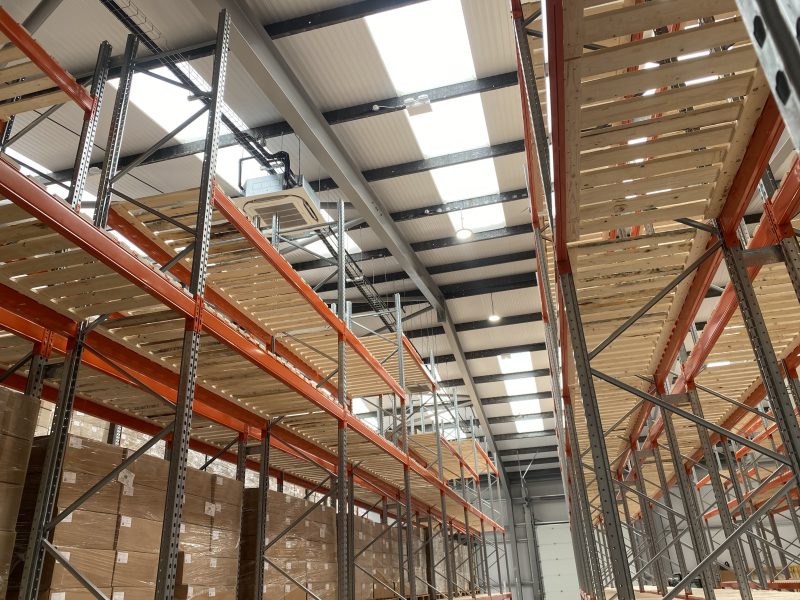 Interior view of a warehouse with tall metal shelving units and wooden platforms, stacked with cardboard boxes under overhead lighting.