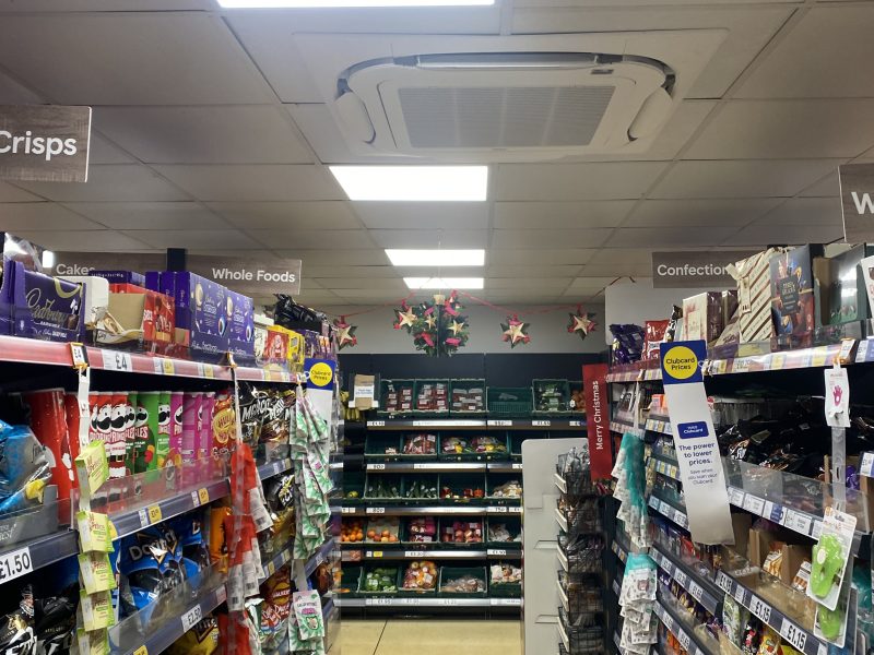 A supermarket aisle with shelves stocked with snacks and other products. Overhead signs read "Whole Foods" and "Confectionery." Bright ceiling lights illuminate the area.