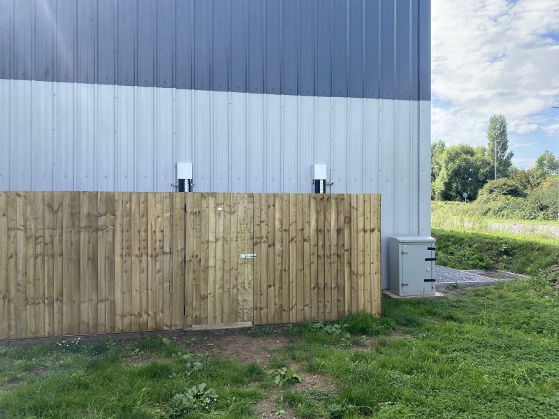 Wooden fence beside a metal building with two mounted devices on the wall and a utility box on the grass.