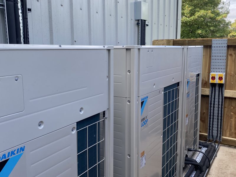 Two large industrial air conditioning units are installed outside a building. They are positioned next to a metal fence and connected to electrical boxes.